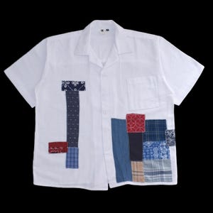 Op de afbeelding: Wit overhemd met korte mouwen en patchworkdetails. Het overhemd heeft een klassieke kraag, een knoopsluiting aan de voorkant en een borstzak. Patches van stoffen met patronen in blauw, rood en beige zijn op de voorkant genaaid.
