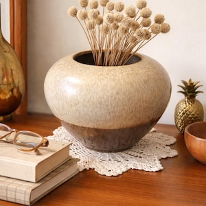 May include: A beige and brown ceramic vase with a round shape, holding dried flower stems. The vase sits on a white lace doily. A gold pineapple and wooden bowl are also visible.