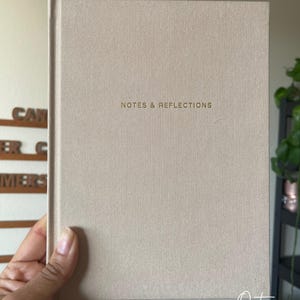 May include: A beige hardcover journal with the gold text "NOTES & REFLECTIONS" centered on the cover. The journal is held by a hand, with a black ribbon bookmark visible. The cover has a textured appearance.