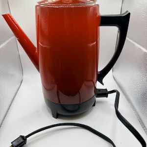 May include: A bright orange electric percolator coffee pot with a black handle, black base, and a clear glass lid knob. The appliance features a long, angled spout and includes a black power cord with a standard two-prong electrical plug.