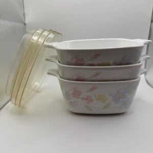 May include: A set of three white square baking dishes with integrated side handles, stacked vertically. Each dish features a delicate floral pattern in soft shades of pink, blue, and yellow. Three clear plastic lids are positioned to the side.