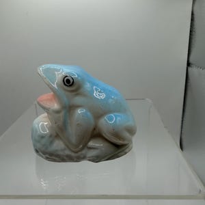 May include: A ceramic frog figurine with a glossy finish, featuring a light blue body, a white underside, and a pale pink interior of the mouth. The frog is depicted in a seated position with its mouth open, showcasing a simple black and white eye.