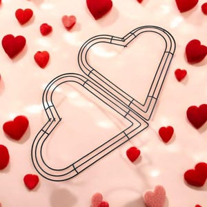 Puede incluir: Un marco de alambre negro en forma de corazón, entrelazado con otro, rodeado de corazones de felpa rojos y rosas de varios tamaños. Los corazones están esparcidos sobre una superficie rosa claro, creando un tema romántico.