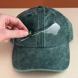 May include: A dark green washed cotton baseball cap featuring an embroidered golf scene on the front. The design includes a white sand bunker, a thin dark green line representing a golf club shaft, and a small red flag atop a white putting green.