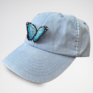 Puede incluir: Una gorra de béisbol de denim azul claro con una mariposa azul bordada en la parte delantera.