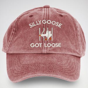 May include: A red vintage-style baseball cap with the text "Silly Goose Got Loose" embroidered in white. A white goose is also embroidered on the cap, appearing to be escaping from a fence.
