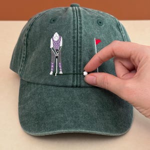May include: A forest green baseball cap featuring an embroidered design of a golfer in a purple argyle vest and grey trousers, standing near a golf flag. A small white ball is attached to the cap by a thin green thread near the golfer.