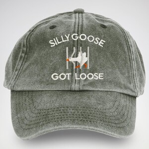 May include: A dark green, distressed, baseball cap with white embroidery that reads "Silly Goose Got Loose". The embroidery also includes a cartoon image of a white goose escaping from a cage.