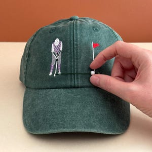 May include: A forest green baseball cap featuring an embroidered design of a golfer in a purple argyle vest and grey pants, positioned next to a golf hole with a red flag. A small white bead is attached to the cap near the golf hole.