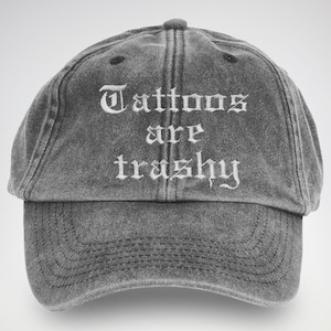 May include: A grey baseball cap featuring the white embroidered words "Tattoos are trashy" in a gothic font. The cap has a curved peak and a faded, vintage appearance.