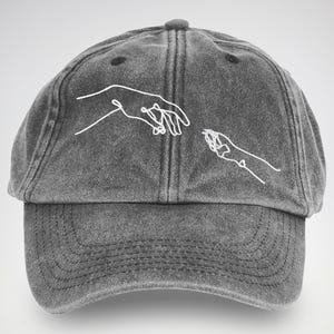 Könnte beinhalten: Graue Baseballkappe mit gebogenem Schirm. Die Kappe hat ein weißes, gesticktes Design von zwei sich fast berührenden Händen. Die Kappe hat einen abgenutzten, Vintage-Look.