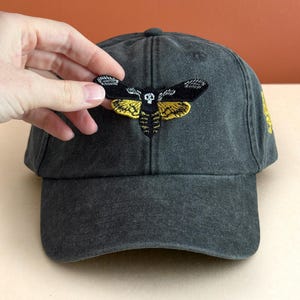 May include: A charcoal gray baseball cap featuring an embroidered patch of a moth with black and yellow wings and a small white skull detail on its thorax. The cap has a structured crown, a curved brim, and a button on the top center.