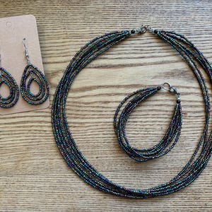 May include: A jewelry set consisting of a multi-strand necklace, a multi-strand bracelet, and a pair of teardrop-shaped earrings on a display card. Each piece is crafted from small, iridescent, dark-colored seed beads with a metallic finish.