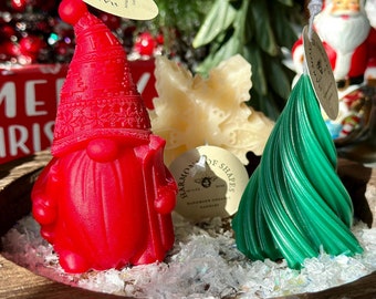 Könnte beinhalten: Drei Weihnachtskerzen in einer Holzschale. Die Kerzen sind in Form eines Zwergs, einer Schneeflocke und eines spiralförmigen Weihnachtsbaums. Der Zwerg ist rot mit einer weißen Mütze und einem weißen Bart. Die Schneeflocke ist weiß und der Weihnachtsbaum ist grün.