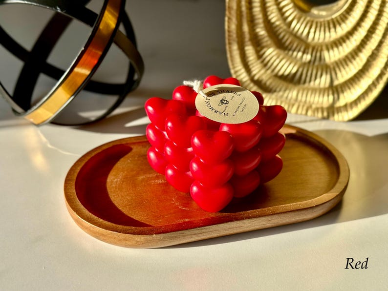 May include: Red heart-shaped candle on a wooden tray. The candle is made by "Harmonia" and has a label that says "Handmade with Love".