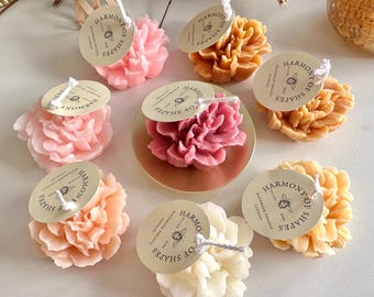 May include: A collection of eight flower-shaped candles in shades of pink, peach, orange, and white. Each candle features a round beige tag printed with the text Harmony of Shapes, a bee illustration, and the words handmade organic candles.