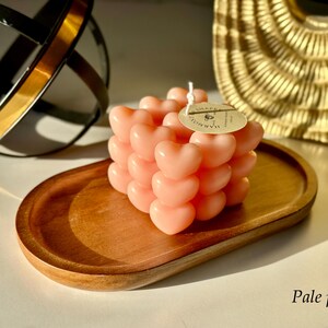 May include: Pale pink heart-shaped candle on a wooden tray. The candle is made of multiple hearts stacked on top of each other. The label on the candle says "remarkable".