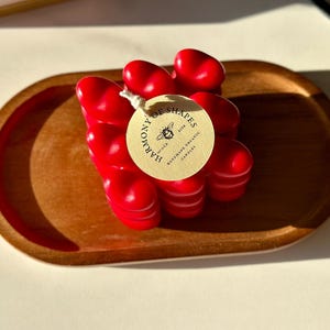 May include: A red heart-shaped candle on a wooden tray. The candle is labeled "Harmony of Shapes" with a bee logo and the text "Hand-Poured Soy & Coconut Candles".