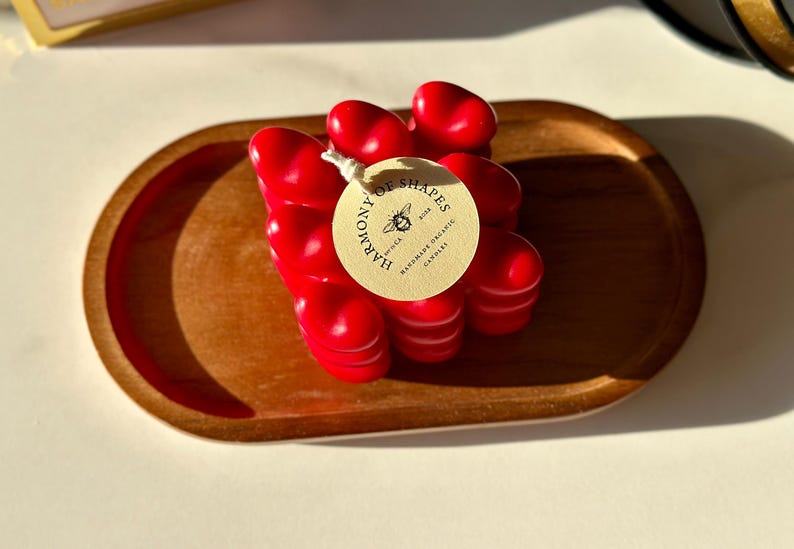 May include: A red heart-shaped candle on a wooden tray. The candle is labeled "Harmony of Shapes" with a bee logo and the text "Hand-Poured Soy & Coconut Candles".