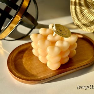 May include: A heart-shaped candle in ivory, arranged in a cube shape, sits on a wooden tray. The candle is uncolored and has a label that reads "Bath & Body Works".