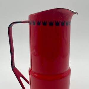 May include: Red enamel pitcher with a black crown design around the top. The pitcher has a black handle.