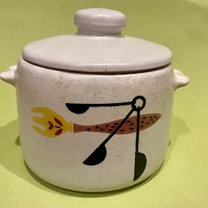 May include: A white ceramic crock with a lid, featuring a brown, yellow, and black design of kitchen utensils. The crock has a speckled texture.