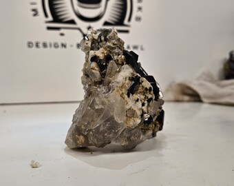 May include: A cluster of clear quartz crystals with black tourmaline inclusions. The crystals are arranged in a pyramid shape and have a rough, natural texture.