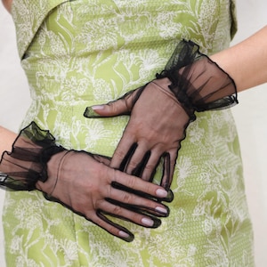 May include: A pair of black sheer gloves with ruffles at the wrist. The gloves are worn by a person wearing a green dress with a white floral pattern.