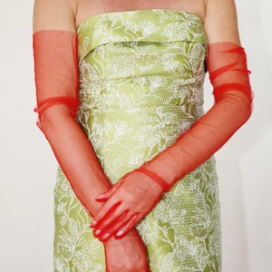 May include: A light green strapless dress with a white floral pattern is worn with long, sheer red gloves. The gloves cover the hands and arms up to above the elbows. The dress has a fitted bodice and a straight skirt.