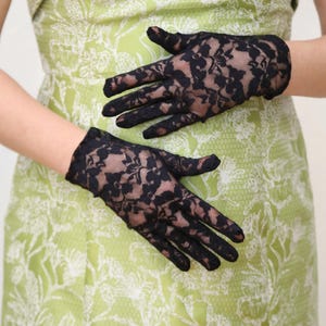 May include: A pair of black lace gloves with a floral pattern. The gloves are worn by a person wearing a green dress with a white floral pattern.