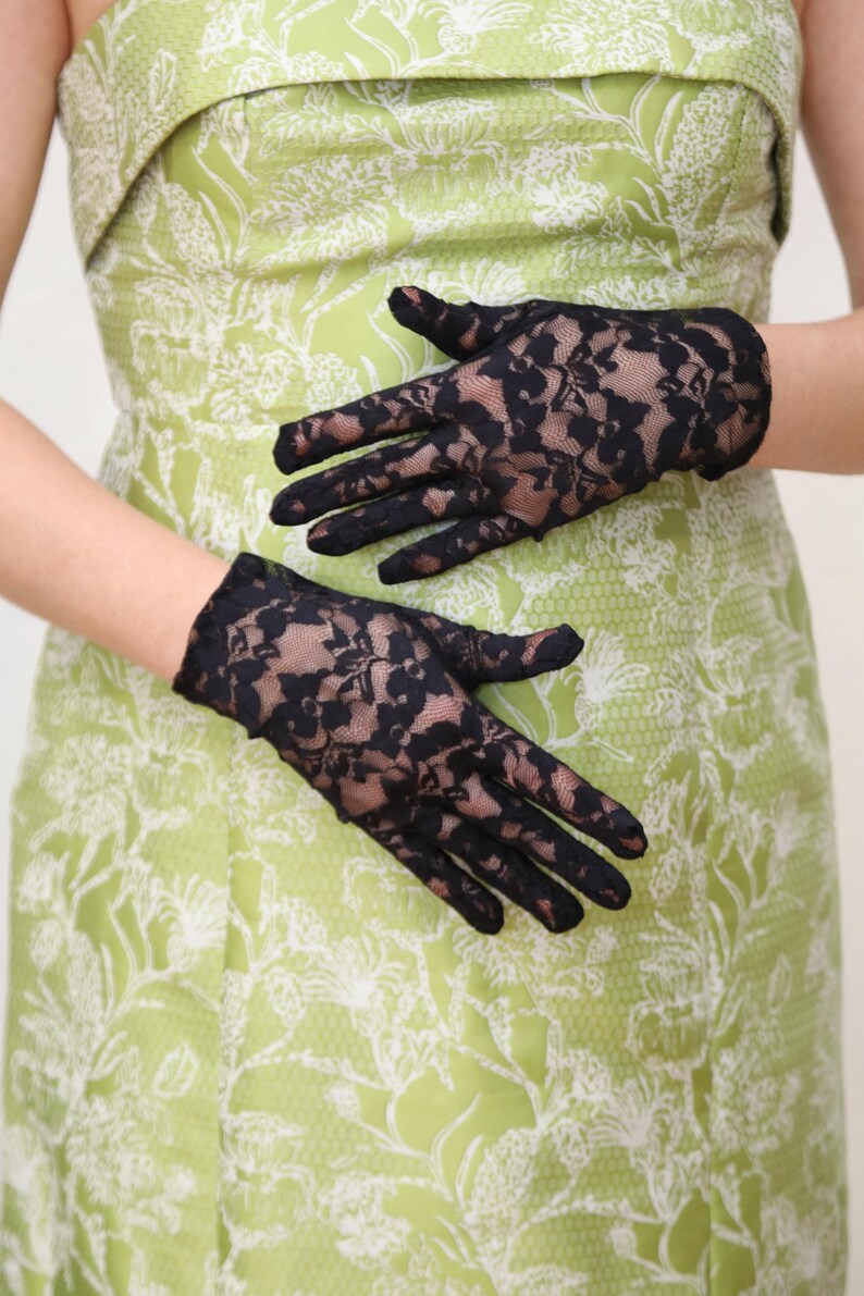 May include: A pair of black lace gloves with a floral pattern. The gloves are worn by a person wearing a green dress with a white floral pattern.