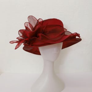 May include: A burgundy hat with a large, ruffled, sheer bow on the side. The hat is on a white mannequin head.