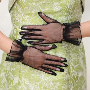 May include: A pair of black sheer gloves with ruffled edges. The gloves are worn by a person wearing a green floral dress.