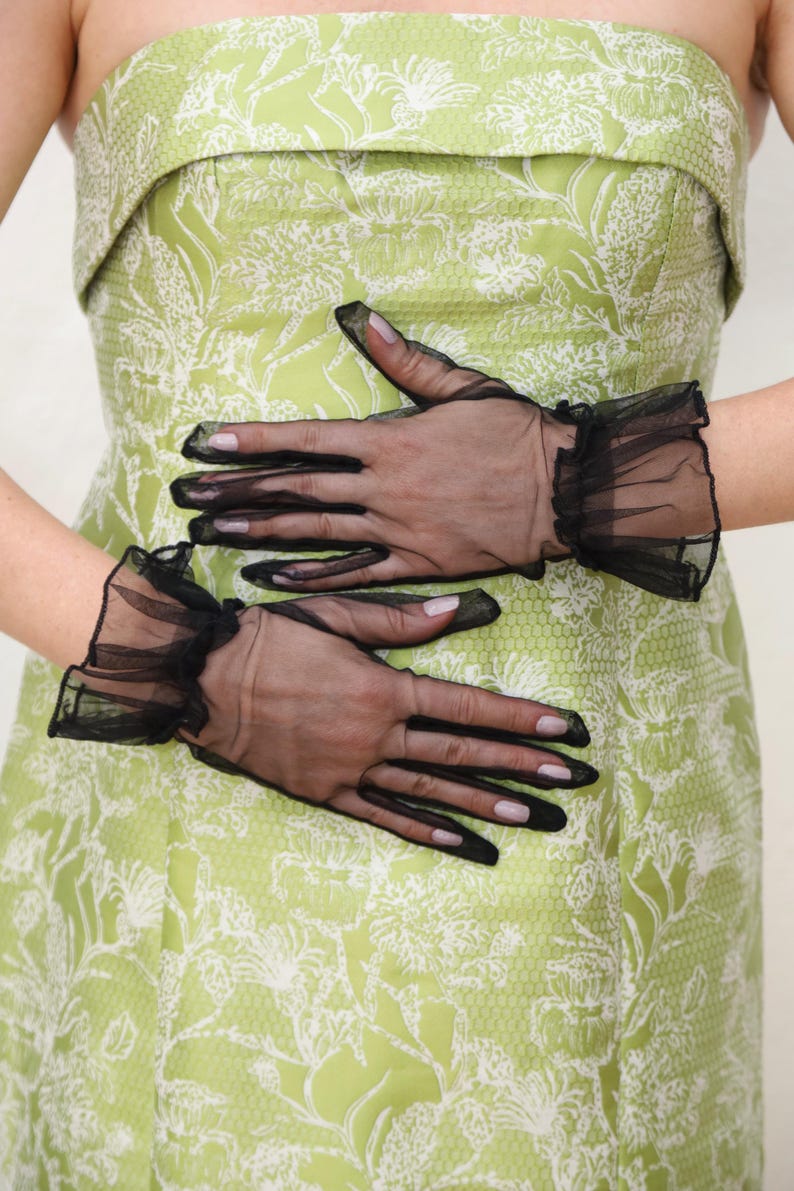 May include: A pair of black sheer gloves with ruffled edges. The gloves are worn by a person wearing a green floral dress.