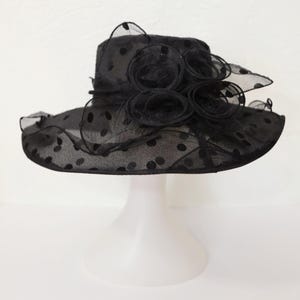 Puede incluir: Un sombrero negro de lunares con un gran adorno de flor negra. El sombrero está hecho de una tela transparente y tiene un ala ancha.