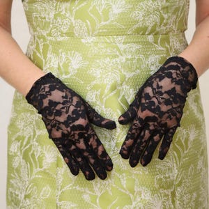 May include: A pair of black lace gloves with a floral pattern. The gloves are worn by a person wearing a green dress with a white floral pattern.