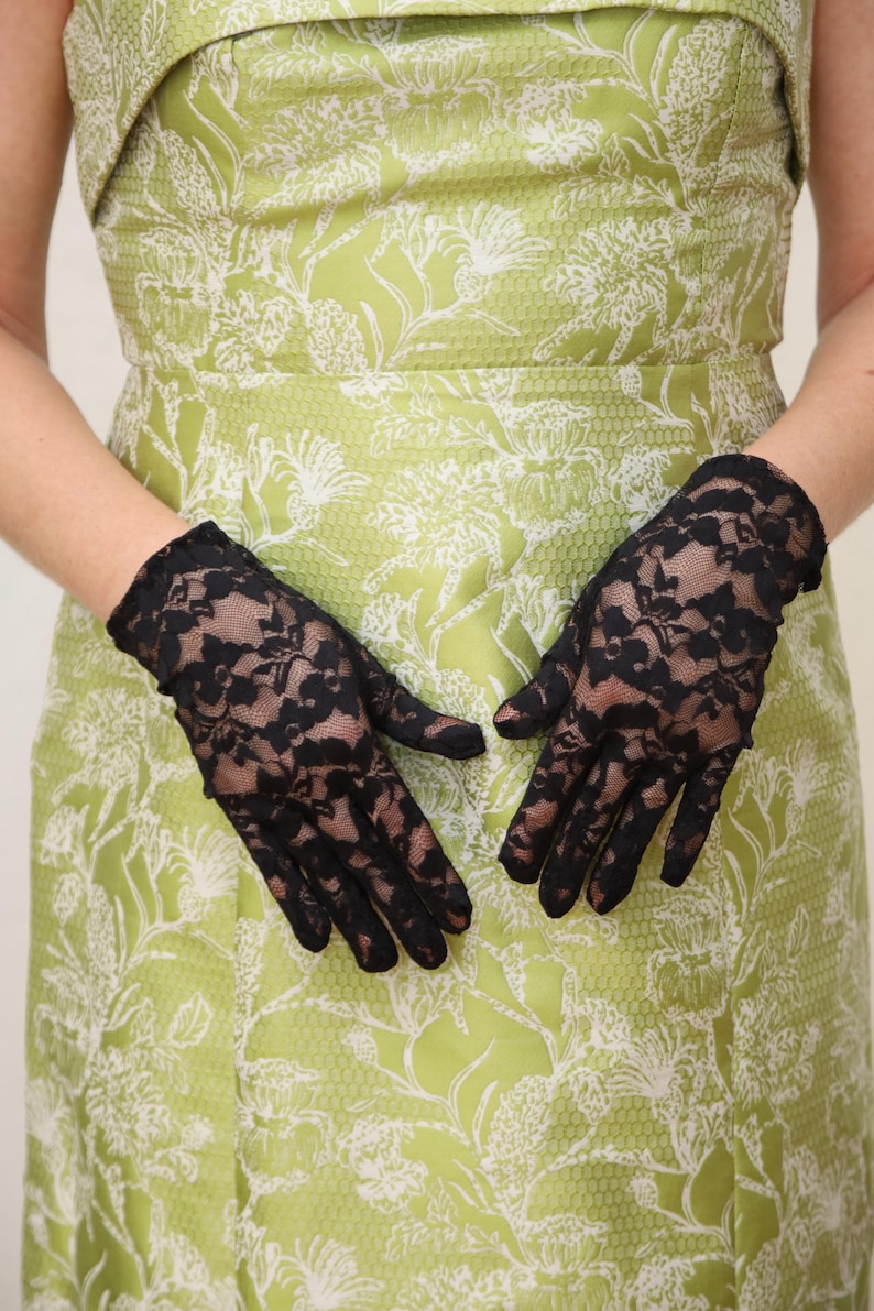 May include: A pair of black lace gloves with a floral pattern. The gloves are worn by a person wearing a green dress with a white floral pattern.