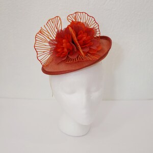 May include: A burnt orange fascinator with a small, round base and a decorative arrangement of orange fabric flowers and fan-shaped accents. The headpiece is displayed on a white mannequin head, showcasing its design.