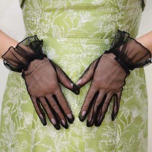 May include: A pair of black sheer gloves with ruffled cuffs. The gloves are worn by a person wearing a green dress with a white floral pattern.