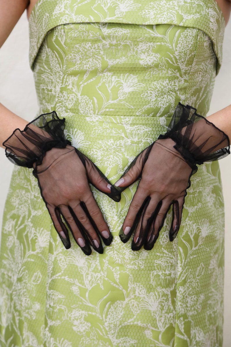 May include: A pair of black sheer gloves with ruffled cuffs. The gloves are worn by a person wearing a green dress with a white floral pattern.