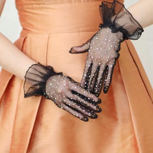 May include: A pair of black sheer gloves with a ruffled cuff and rhinestones. The gloves are worn on a person's hands.