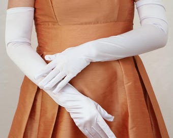 May include: White satin opera gloves, reaching the upper arm, are crossed over a copper-colored sleeveless dress. The dress has a fitted bodice and a full skirt, creating an elegant silhouette. The gloves add a touch of sophistication.