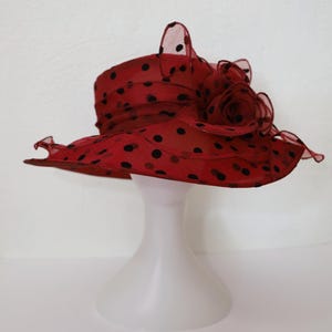 Puede incluir: Un sombrero rojo con lunares blancos y una gran flor negra en un lado. El sombrero está hecho de una tela transparente y tiene un ala ancha.