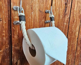 May include: A nautical-themed toilet paper holder. The holder features a thick, off-white rope threaded through two chrome-colored metal fixtures. The rope supports a roll of white toilet paper against a wooden background.