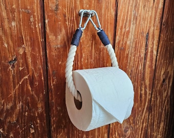 May include: A nautical-themed toilet paper holder. The holder features a thick, off-white rope with navy blue accents, suspended by a metal bracket. A roll of white toilet paper is held within the rope loop, set against a rustic wooden door.