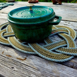May include: A teal green cast iron pot with a lid, resting on a handmade rope trivet. The trivet is made of thick, braided rope in shades of tan and blue. The pot has a gold-colored handle on the lid.