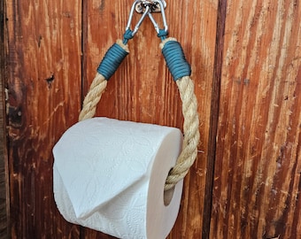 May include: A rustic toilet paper holder made of thick, natural rope and teal-colored wrapped accents. The rope is attached to a metal bracket with two carabiner-style clips. A roll of white toilet paper is in the holder, ready for use.