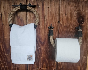 May include: A rustic bathroom set featuring a towel ring and a toilet paper holder. The towel ring is made of rope and black metal, holding a white towel. The toilet paper holder is also made of rope and black metal, holding a roll of white toilet paper.
