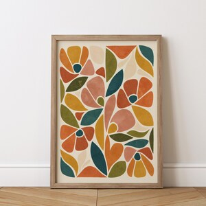 May include: A framed art print featuring a floral design with abstract shapes in orange, yellow, pink, and teal. The artwork is set against a cream background and framed in a light wood. The print is displayed on a wooden floor.