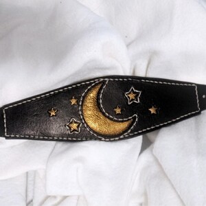 May include: Black leather headband with a gold crescent moon and gold stars. The celestial design is accented with white stitching. A unique accessory for a celestial or bohemian style. The headband is approximately 20 cm long.